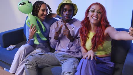 studio shot of group of gen z friends sitting on sofa posing for selfie with toy alien on mobile phone