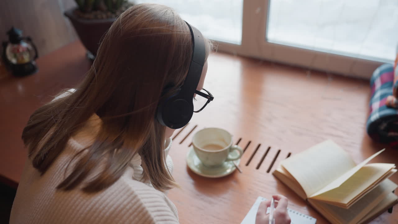 rear view of student with headphone writing in notebook from open textbook on wooden table with ceramic mug and folded cloth in background