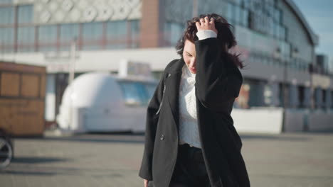 young woman scratches her hair while looking left, wearing white cable knit sweater and black coat, on sunny urban street with modern buildings and parked green bicycles in background