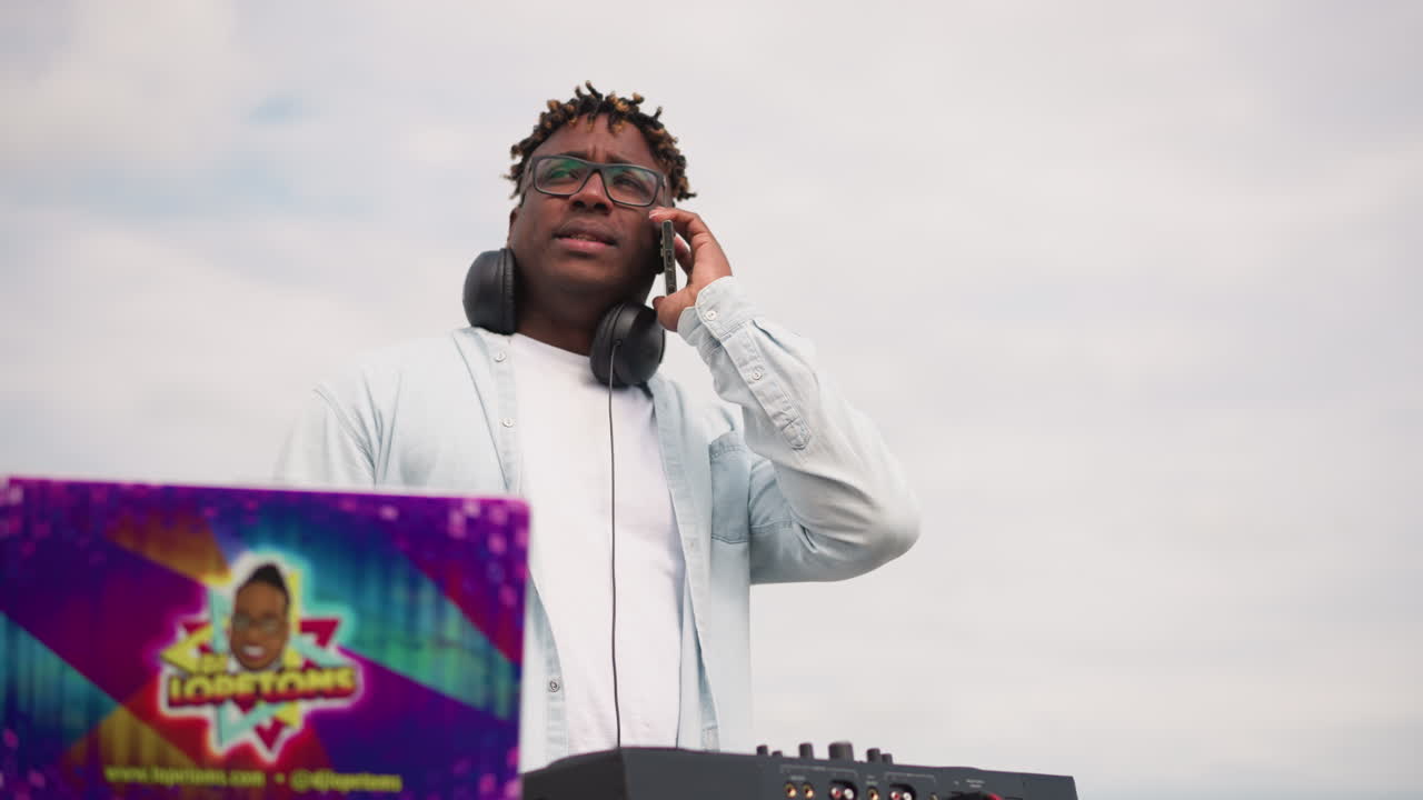 DJ nero che mixa su un laptop colorato, cuffie al collo, multitasking con una telefonata, giradischi e controller su un ponte sul tetto, vivaci adesivi artistici, energia vivace da festa, atmosfera da festival.