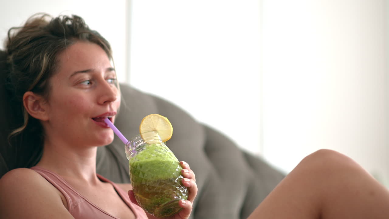Primo piano di una donna che beve un succo verde mentre è seduta su un divano