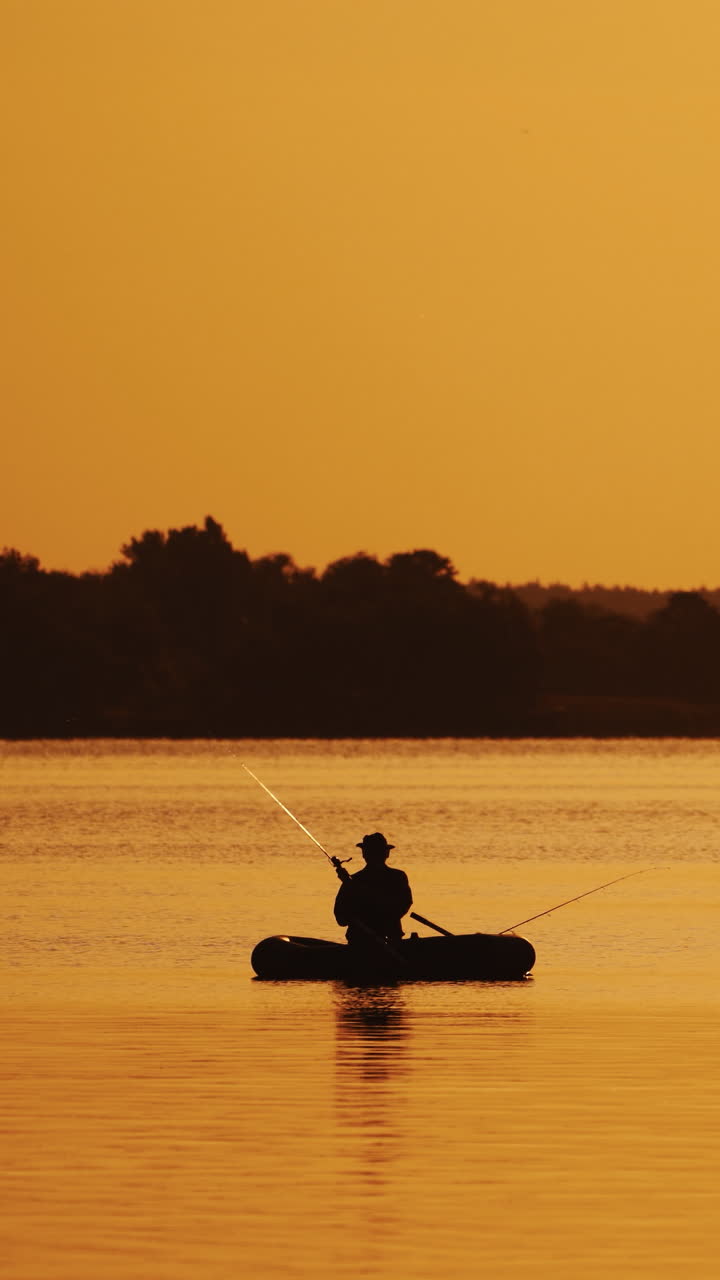 Amazing view of the evening sunset and a man in a boat fishing. Silhouette of a fisherman sitting in a row boat and catching fish on the golden evening background over the river surface. Vertical video