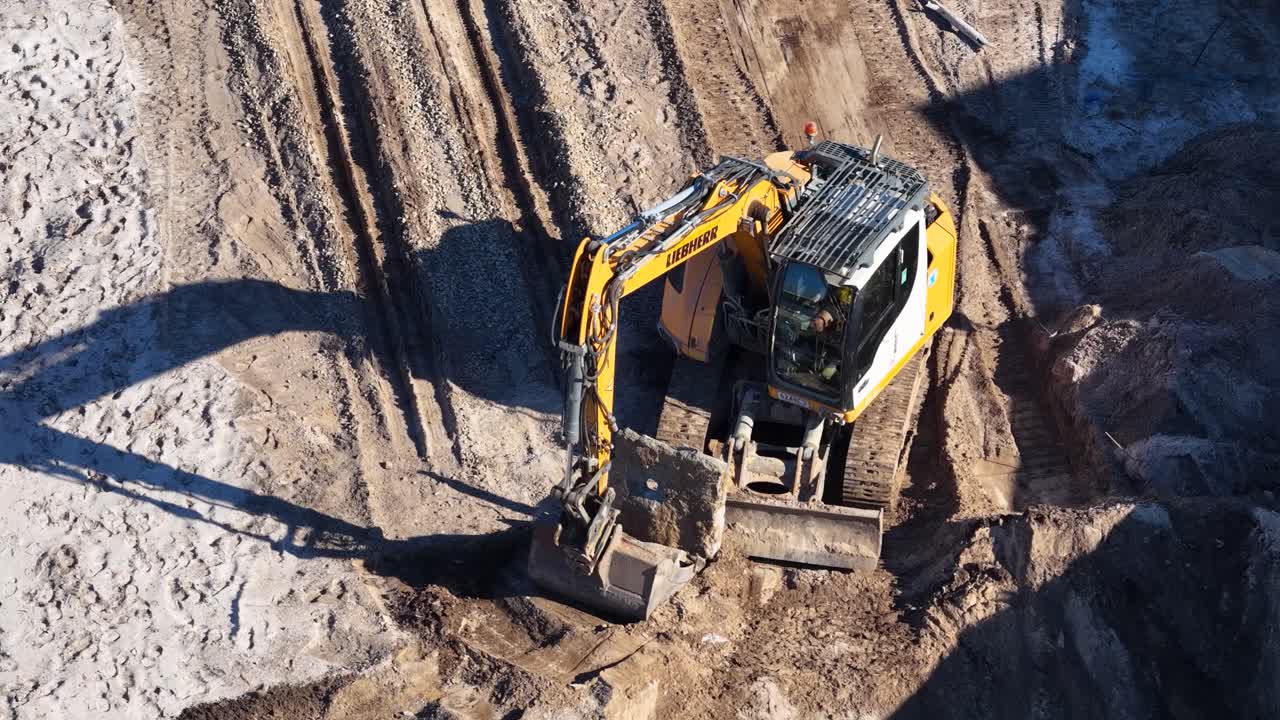 Yellow excavator operates on dirt, digging and moving soil, morning sunlight, aerial perspective