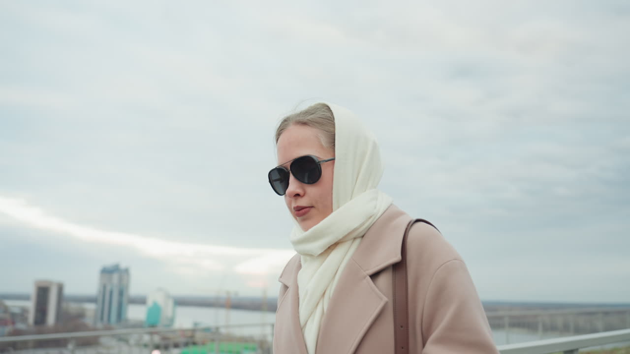 Donna caucasica che controlla il telefono durante il tragitto mattutino su un tetto, scorrendo playlist e messaggi, preparandosi a indossare cuffie over-ear, sistemandosi sciarpa e occhiali da sole, con lo skyline urbano sullo sfondo, energia da content creator.