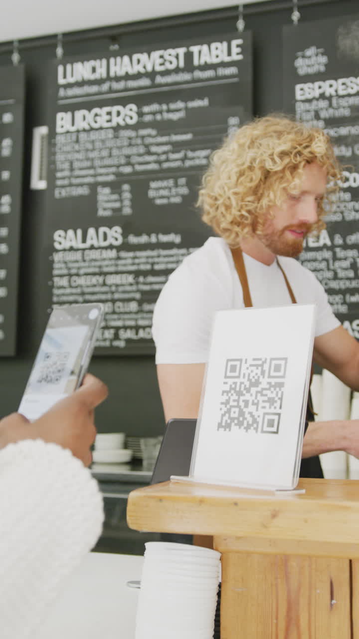 Vertical video of happy diverse male barista and woman scanning qr code with smartphone in cafe