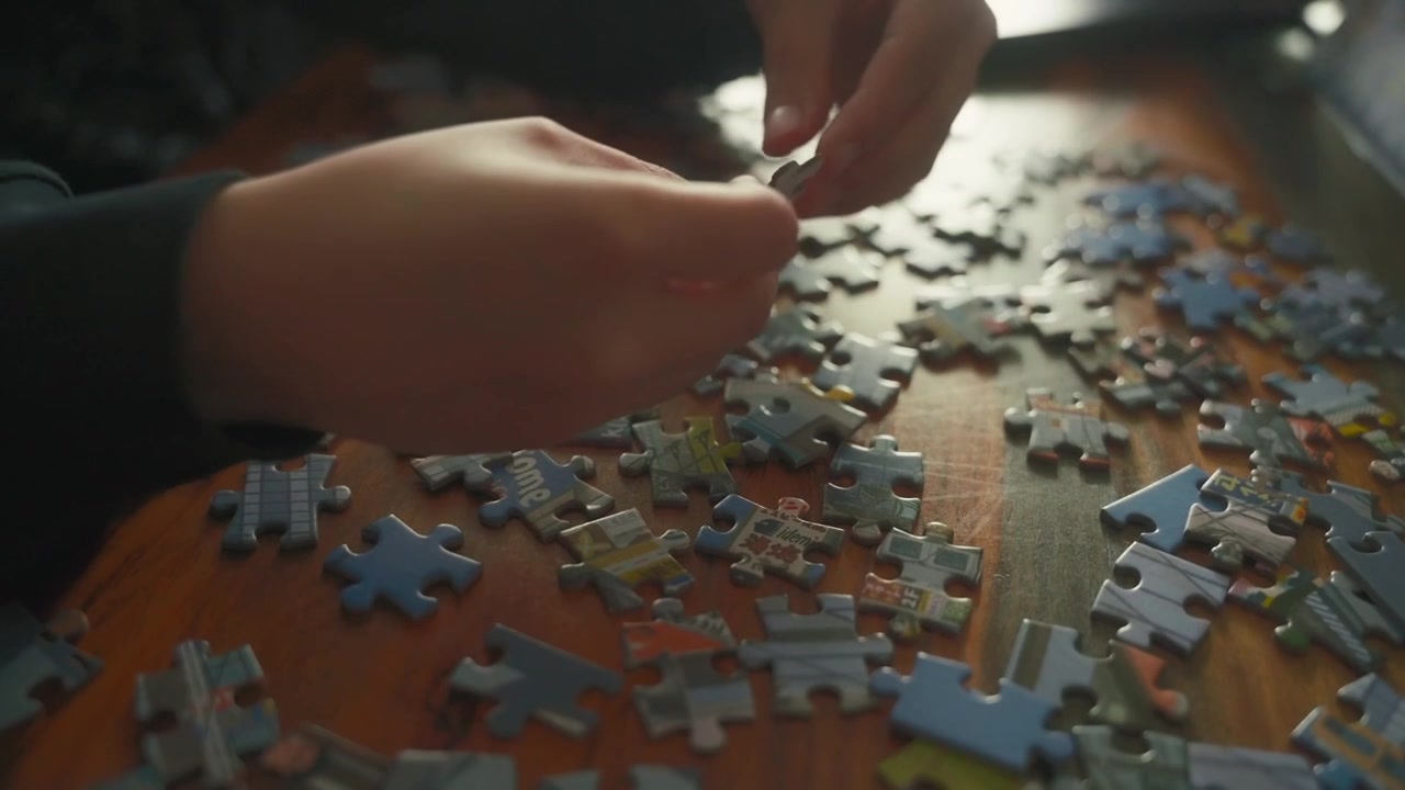 A steady close-up shows hands picking up and sorting jigsaw pieces on a wooden table. Soft light creates a focused, quiet scene for hobby, problem-solving and leisure content.