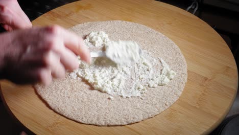 Un-Primer-Plano-De-Un-Pan-De-Tortilla-En-Una-Mesa-De-Madera,-Mientras-Un-Chef-Esparce-Una-Cucharada-De-Queso-Bajo-En-Grasa-De-Manera-Uniforme-Antes-De-Agregar-Una-Porción-De-Relleno-De-Pollo-Picante-Y-Doblar-La-Envoltura