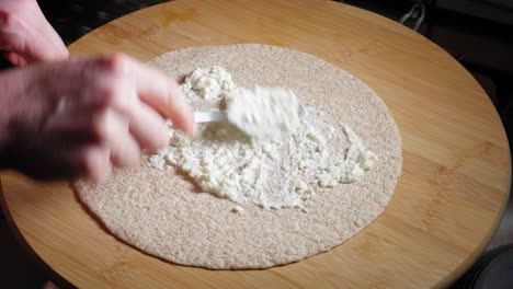un primer plano de un pan de tortilla en una mesa de madera, mientras un chef esparce una cucharada de queso bajo en grasa de manera uniforme antes de agregar una porción de relleno de pollo picante y doblar la envoltura