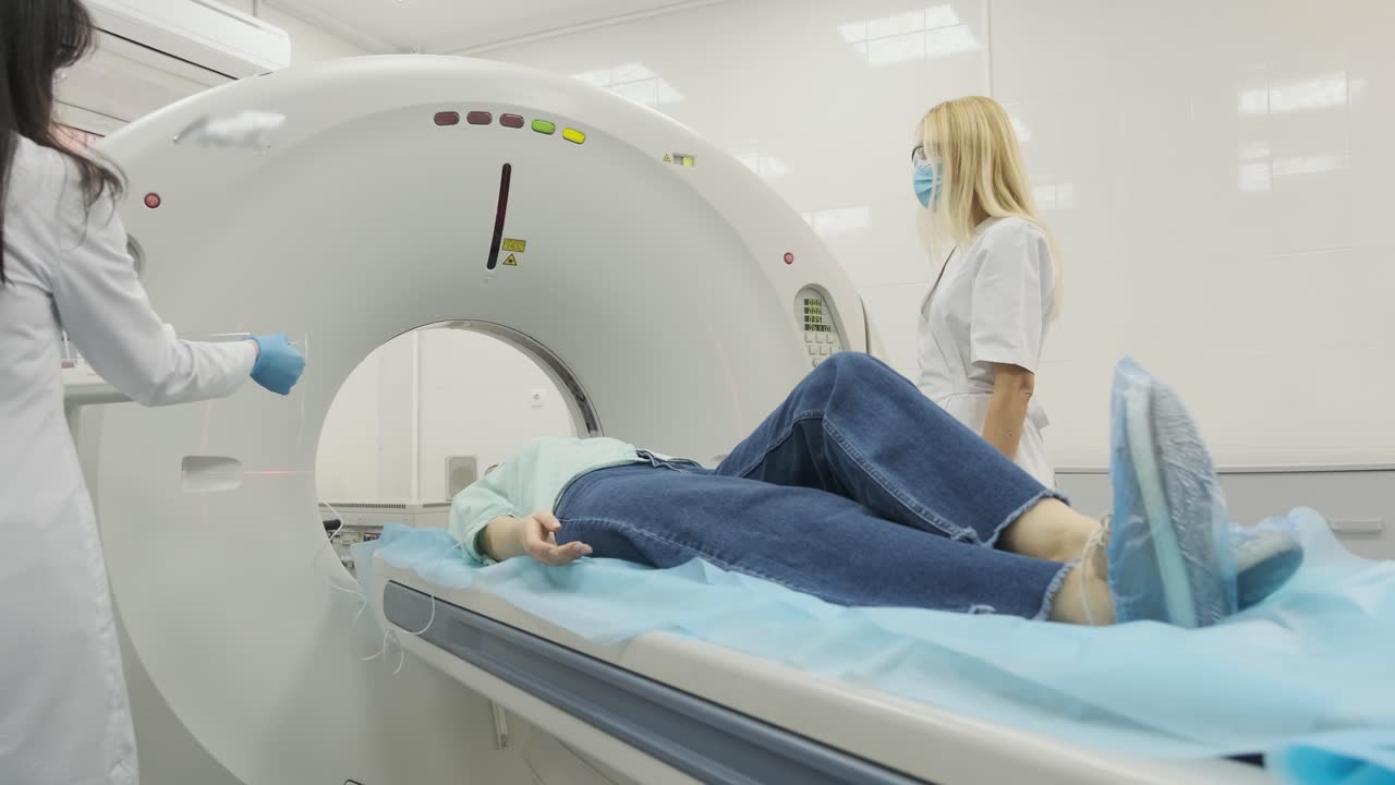 la paciente está siendo sometida a una tomografía computarizada o resonancia magnética bajo la supervisión de dos radiólogos calificados en una clínica médica moderna. la paciente está acostada en una mesa de tomografías computarizadas o mri, moviéndose fuera de la máquina