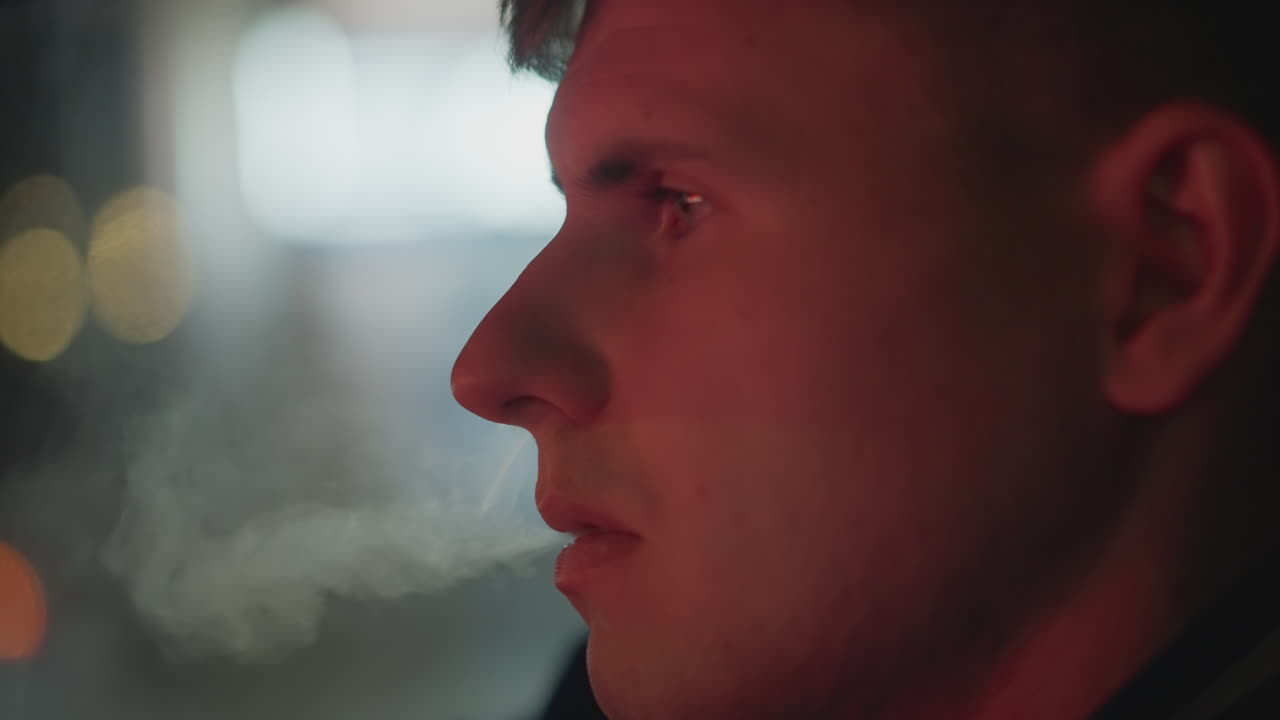 Nighttime urban driving, Driver exhales smoke behind foggy glass illuminated by city lights, Lone driver with tense jaw exhales cigarette smoke through fogged windshield at night in city