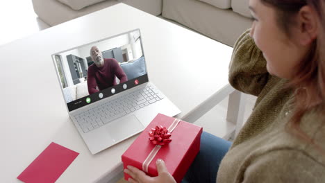 Caucasian-woman-holding-red-gift-using-laptop-with-african-american-man-on-screen