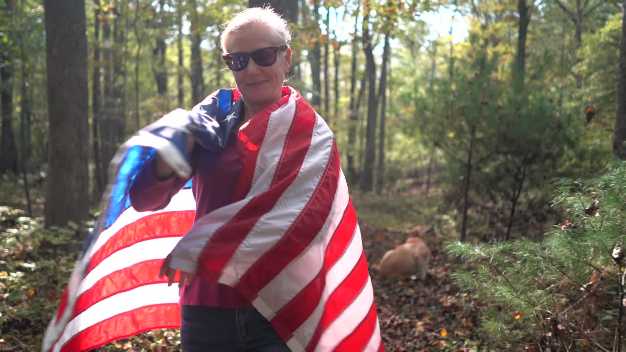 Smilende, blond kvinde, der går gennem skoven og vikler et amerikansk flag om sig