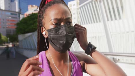 african american woman in face mask wearing earphones while standing on the city bridge