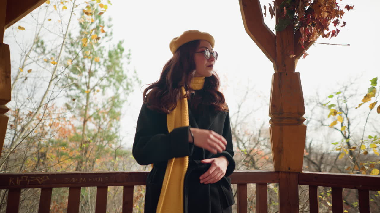 En elegant dame med gul beret og skjerf hviler på et lysthusrekkverk med hendene knyttet sammen, justerer håret mens hun ser seg rundt i et høstlig skogsmiljø.