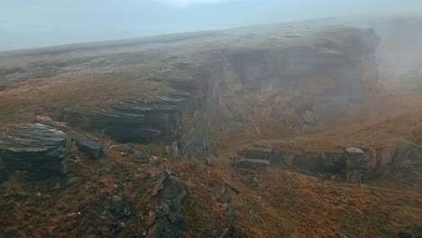 Dramatic-aerial-view-of-clouds-moving-slowly-over-the-hills,-Pennines-on-a-foggy-morning,-golden-hills-and-beautiful-rocky-cliffs-and-moorlands