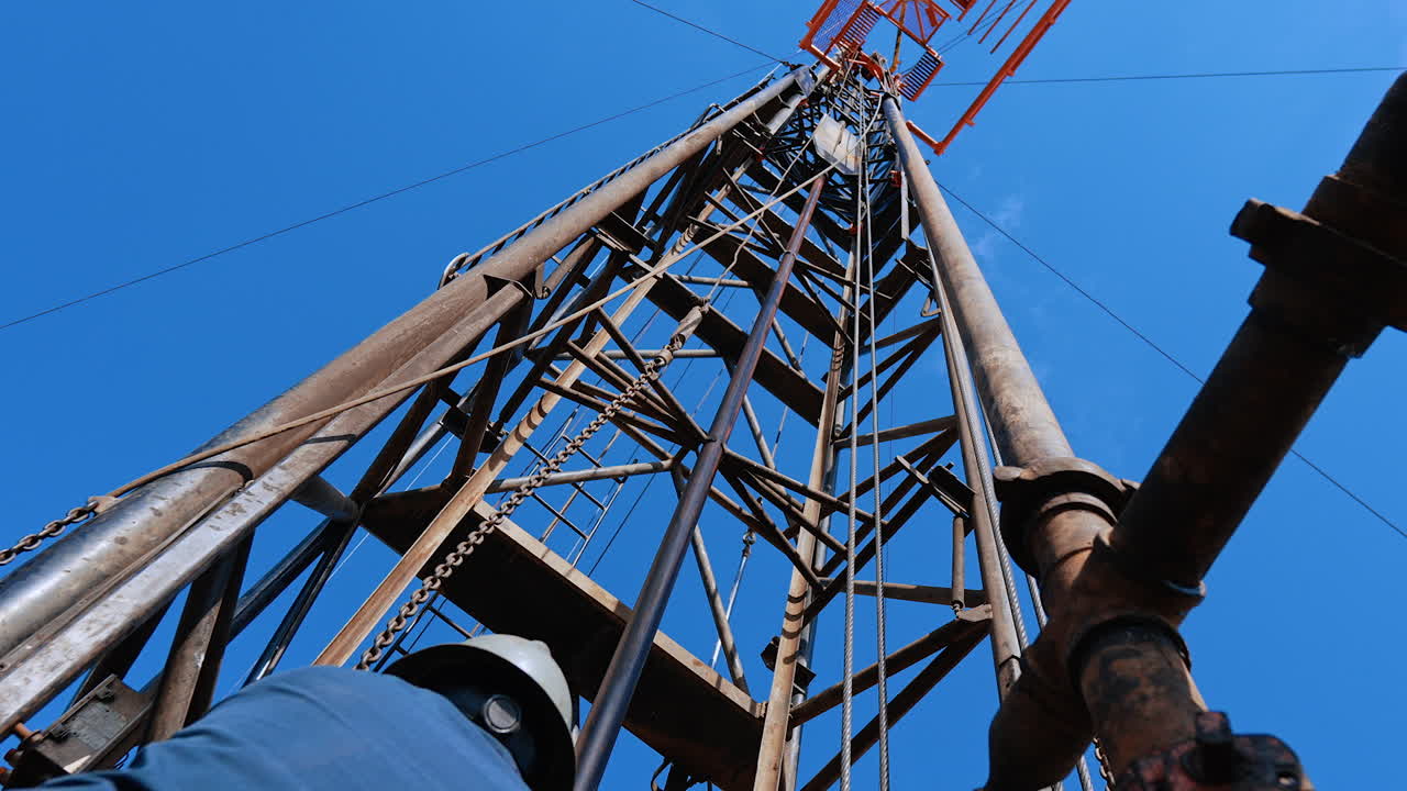 Man in blue uniform and helmet standing at the drilling tower pulling the chain. Worker setting equipment for boring oil or gas.