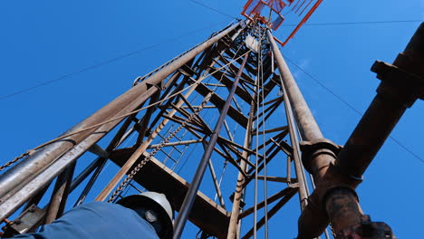 man in blue uniform and helmet standing at the drilling tower pulling the chain. worker setting equipment for boring oil or gas.
