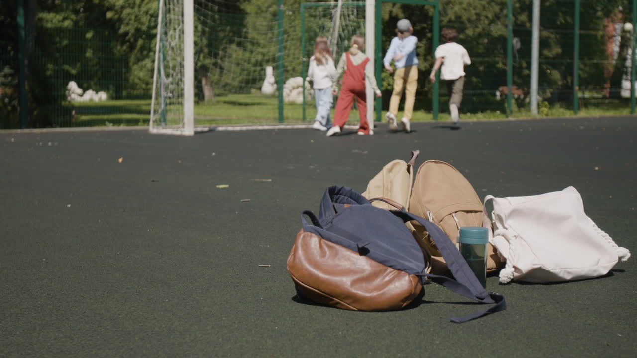 Children playing soccer on an outdoor court with backpacks in the foreground