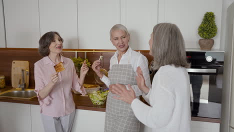 group of cheerful senior friends laughing and toasting with glasses of wine in the kitchen