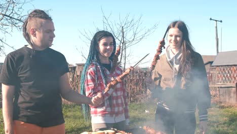 group of young people friends barbecue shashlik meat on top of charcoal grill on backyard. talking and smiling together.