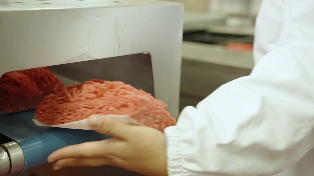 Factory worker in a white coat lifts shaped mince blocks from a conveyor as they exit an automated forming machine.