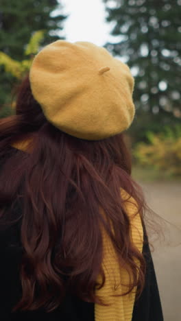 vista trasera de una artista con el cabello rojo largo y ondulado que lleva un abrigo negro, una boina amarilla y una bufanda a juego, ajustando su cabello mientras camina a través de un exuberante parque verde adornado con coloridos follajes de otoño