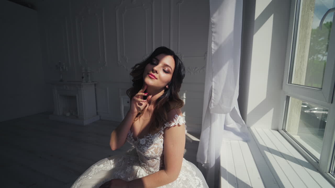Young bride in wedding dress. Luxuty model looking at mirror sitting on chair like at home in studio room with big window