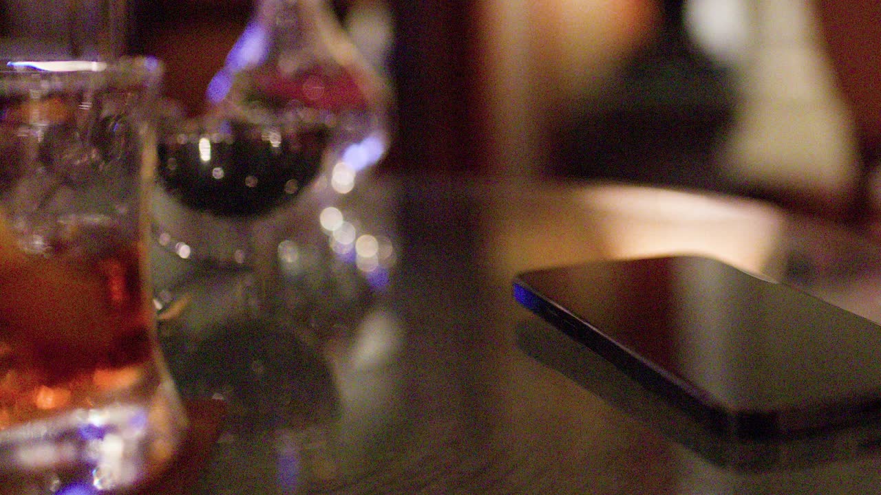 Close-up of whiskey glass with ice on reflective bar table, warm ambient lighting, shallow focus