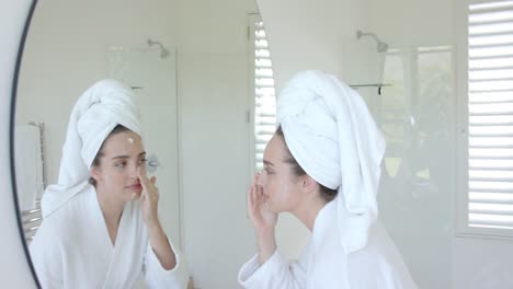 caucasian woman with towel on head applying cream on face in bathroom in slow motion