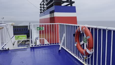 White-railing-of-a-ferry-with-a-lifebuoy-and-stairs,-a-chimney-in-the-background-and-the-wide-sea