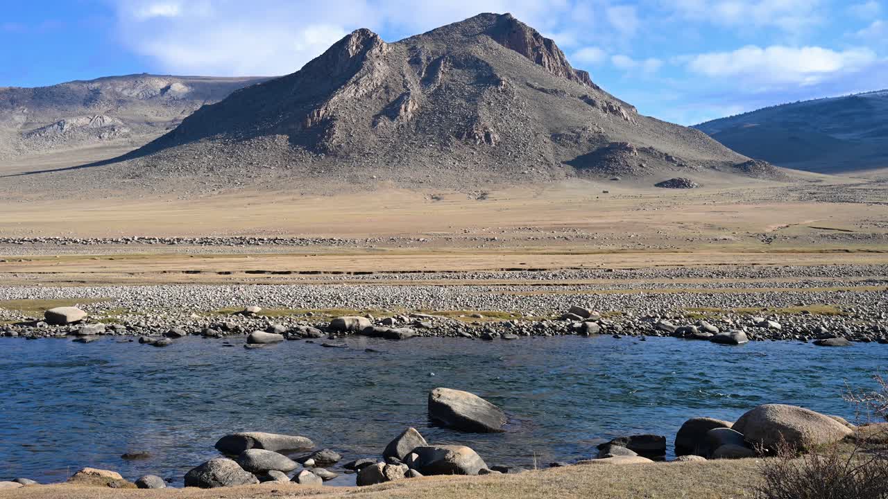 A serene river reflects a majestic, rugged mountain in a vast and remote Mongolian landscape. This tranquil autumn scene showcases the pristine water flowing through the arid, untouched steppe