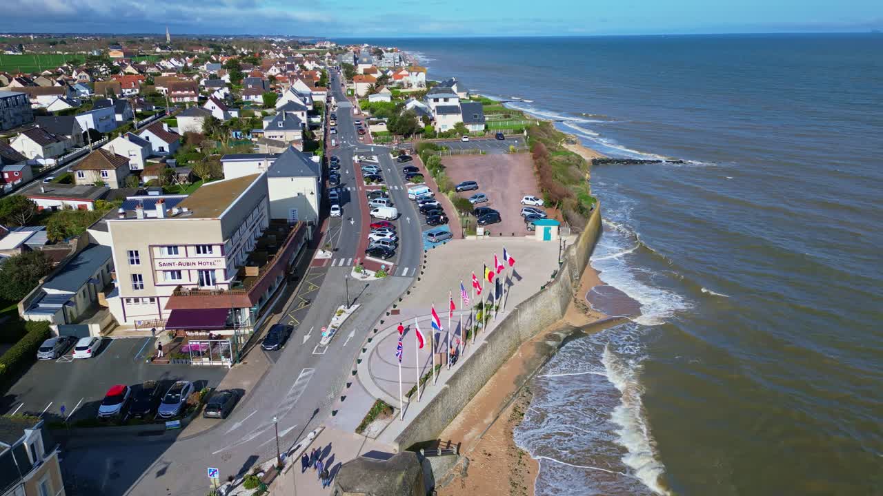 Saint-Aubin-sur-Mer coastline, beach, sea wall promenade with flags, and town, Normandy, France. Aerial forward
