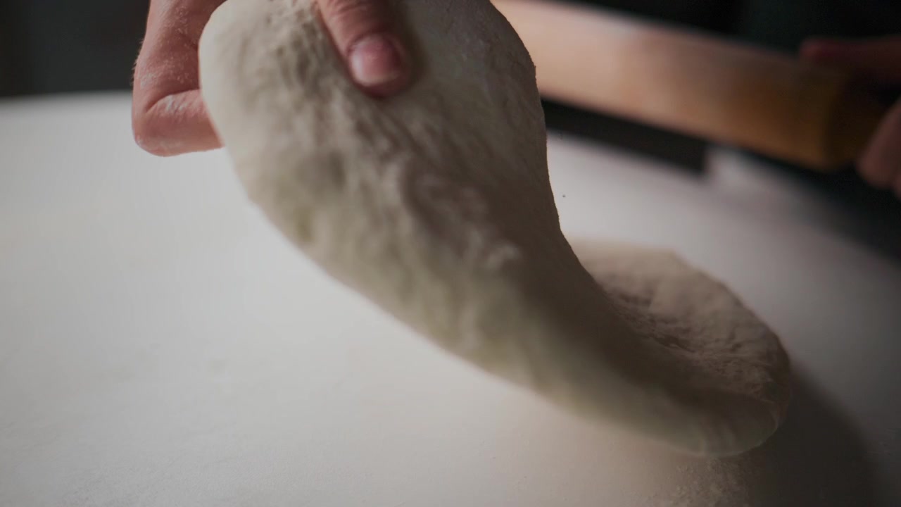 Close-up tangan menggulung adonan dengan penggiling kayu di permukaan putih. Jari-jari menekan adonan dengan lembut, menciptakan tekstur yang halus. Adonan tampak licin dan sedikit elastis. Orang tersebut terlihat fokus dan tenang.