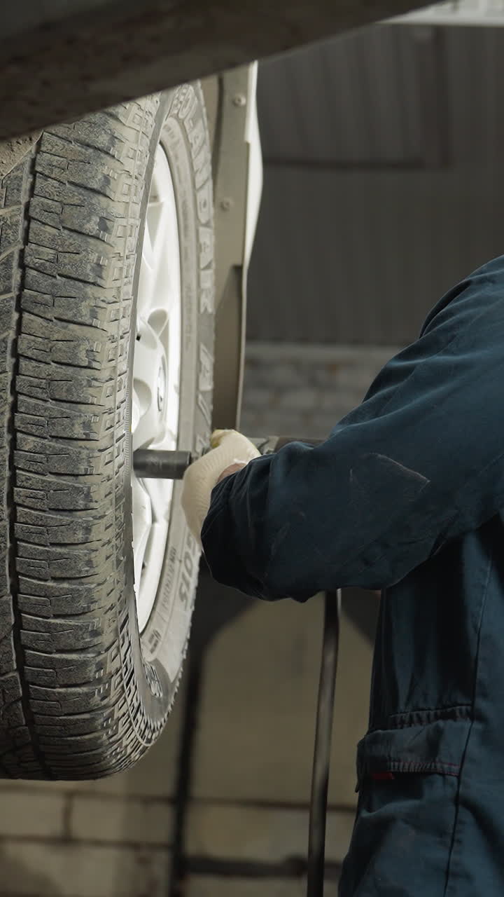 vista aérea de un mecánico en uniforme azul usando una herramienta neumática para apretar los pernos de las ruedas de un automóvil levantado en el taller, mientras que el compañero se acerca, observa el trabajo y asume en aprobación