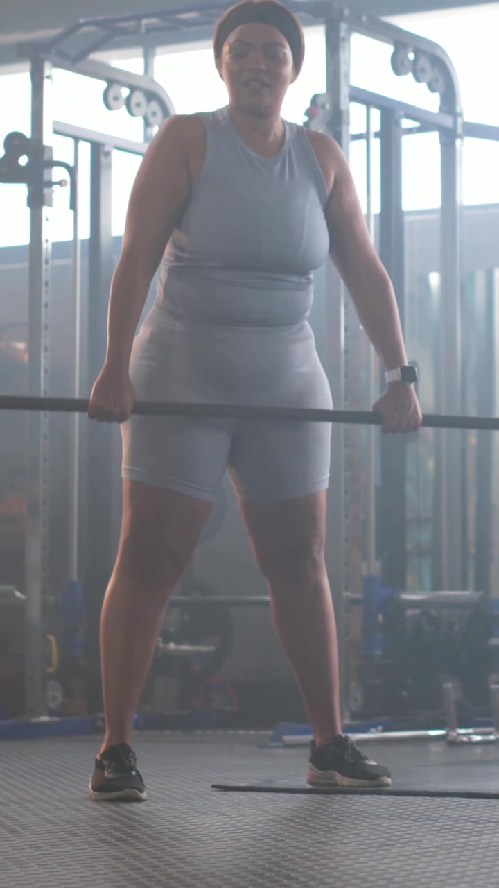 Video verticale: Donna che regola la presa, piegando i fianchi e sollevando un bilanciere in palestra per la tecnica, con un top chiaro e pantaloncini.