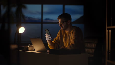 tired young man working on a laptop late night in the office. sleepy businessman sitting at desk in dark office. tired and stressed businessman in glasses works on a laptop of the night city office