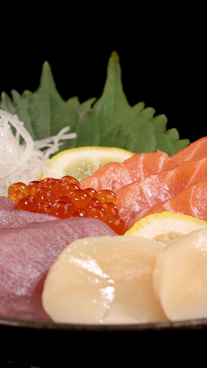 Uma tomada rotativa em close-up de um prato de sashimi japonês com salmão, atum, vieira, ovas de salmão, limão e verduras em um fundo preto sob iluminação de estúdio