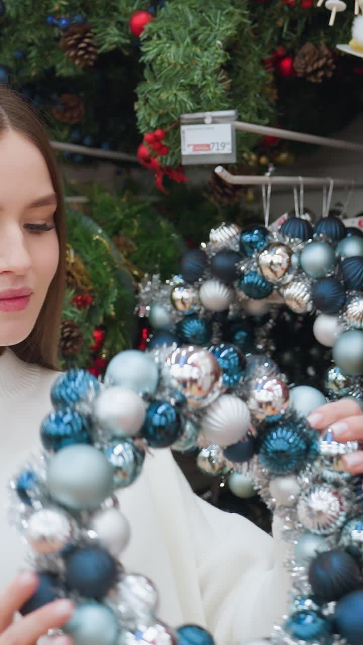 Young woman in white sweater picks up a beautiful Christmas decor wreath and playfully places it on her head while shopping in a festive holiday store surrounded by colorful ornaments