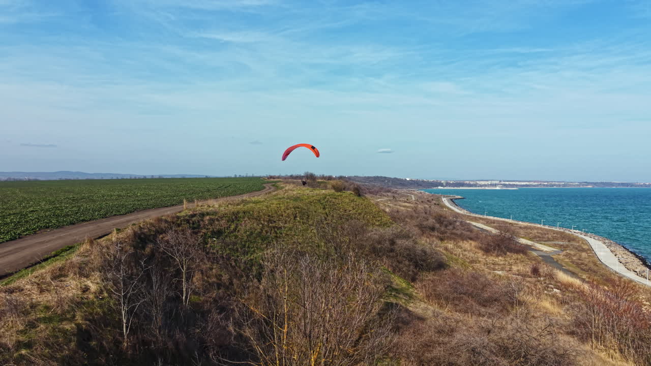 Paraglider soars over a coastal path near a grassy hill with clear skies and a view of the sea on a sunny day