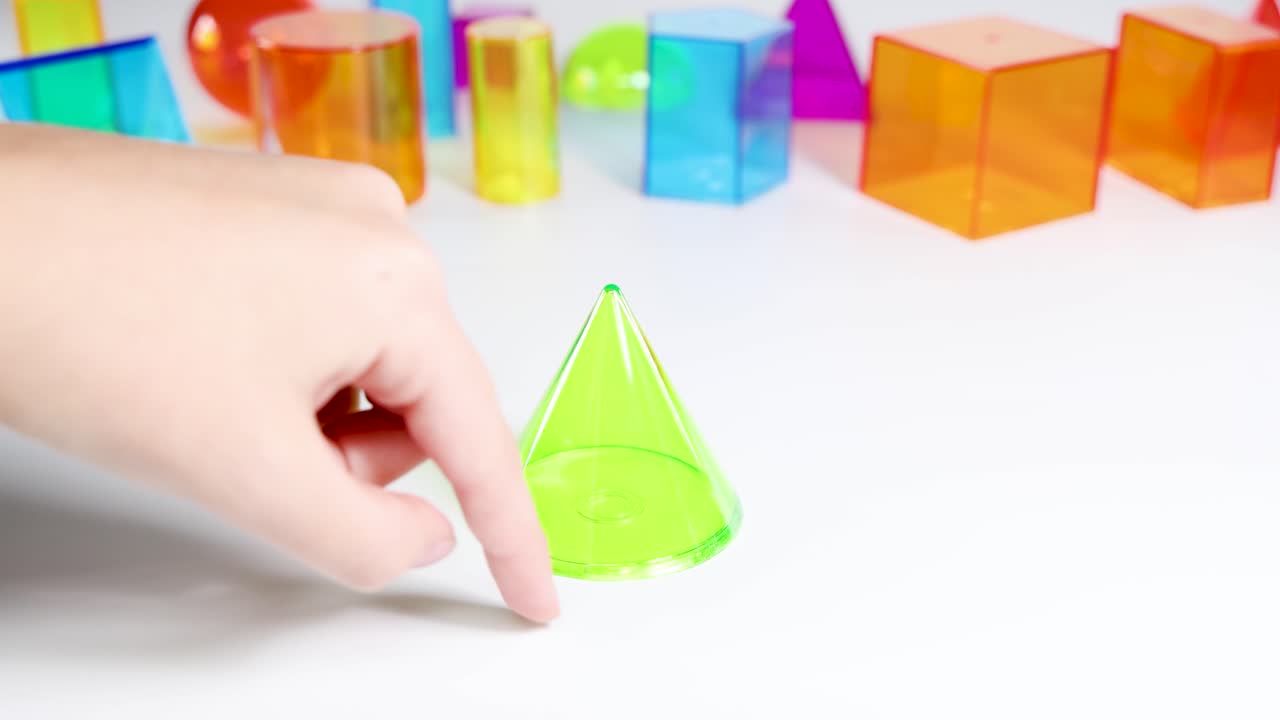 Hands manipulate vibrant 3D shapes on a white surface, highlighting geometry and color in a playful educational setting