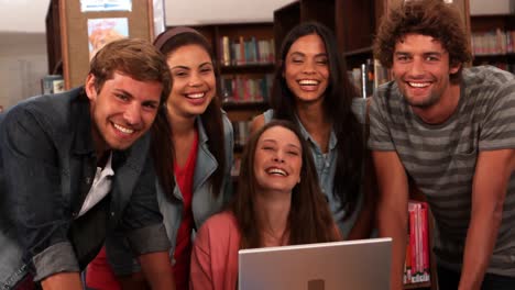 Estudiantes-Felices-De-Pie-En-La-Biblioteca-Con-Una-Computadora-Portátil