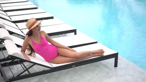 Mujer-Asiática-En-Forma-Sexy-Acostada-Junto-A-La-Piscina-En-Una-Tumbona-Con-Un-Traje-De-Baño-Monokini-Rosa,-Gafas-De-Sol-Y-Un-Sombrero-Posando-Para-El-Fotógrafo