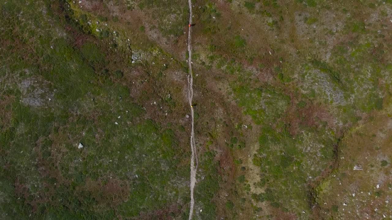 Top down aerial view from high altitude following two hikers or backpackers walking or hiking on a rural Sweden countryside muddy road in the middle of grassy green and brown landscape, cloudy daytime