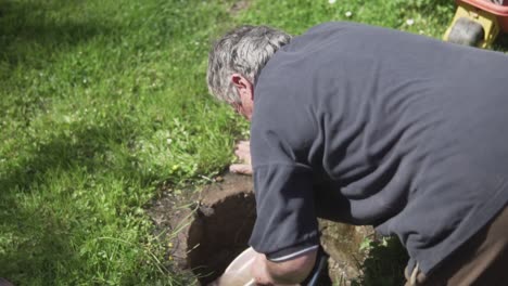 slow motion back shot of old man kneeling and pulling out water from a ground hole using a bucket