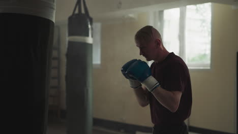 boxer fights punching bag in empty gym. strong man in boxing gloves punches hanging bag in fighting club slow motion. martial arts master