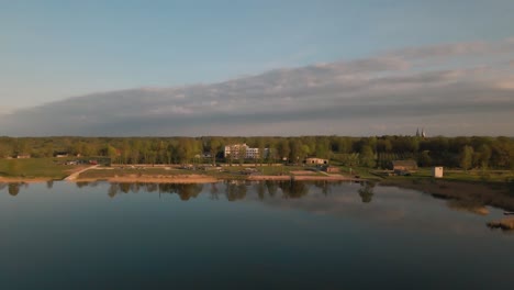 drone footage flying backwards over a calm lake with mirror-like reflections. a small crowd gathers by a lakeside stage adding a serene atmosphere to the scenic landscape at sunset.