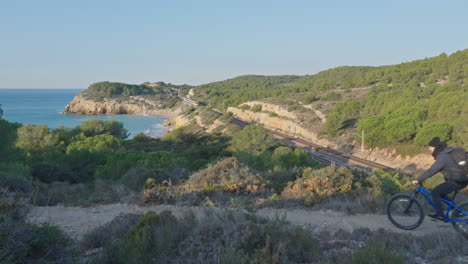 a train goes through mountains while a cyclist rides a bike on a dirt road during early sunny morning