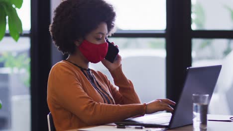 mujer de negocios de raza mixta con máscara sentada usando una computadora portátil en la oficina