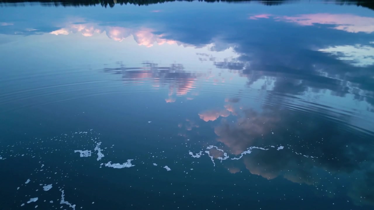 Showing calm lake mirroring pink clouds at dusk foam shifting treeline breeze stirring ripples