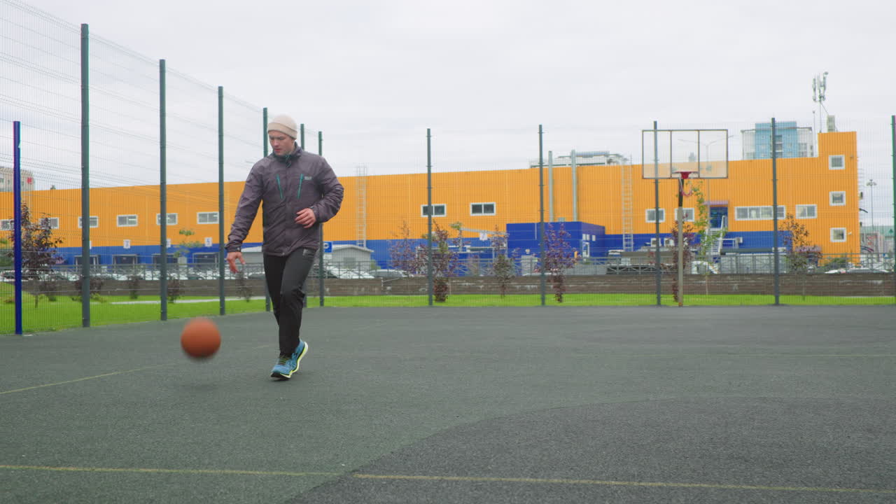 uomo che palleggia con un pallone da basket su un campo all'aperto, indossa un berretto e una giacca, si esercita nel layup e nel tiro in sospensione sotto un cielo nuvoloso, parco giochi urbano recintato con edificio industriale giallo sullo sfondo, grintoso